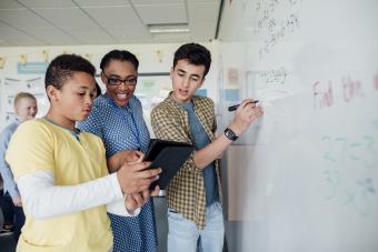 teacher assists two students in a math class at a white board while looking at a tablet